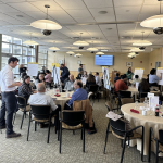 A networking session between participants in a cafe style setting with circular tables, chairs, and large easel notepads